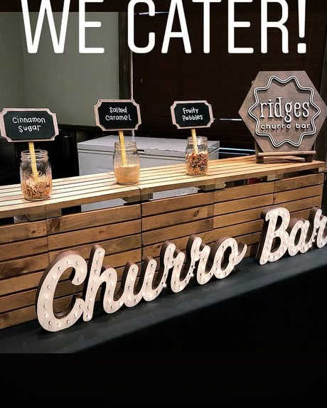 Ridges 'We cater!' setup — a wood-plank churro bar with chalkboard topping signs and a lit 'Churro Bar' marquee.