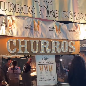 The lit Ridges Churro Bar stall at night with its 'Churros + Ice Cream' banner and marquee CHURROS letters.